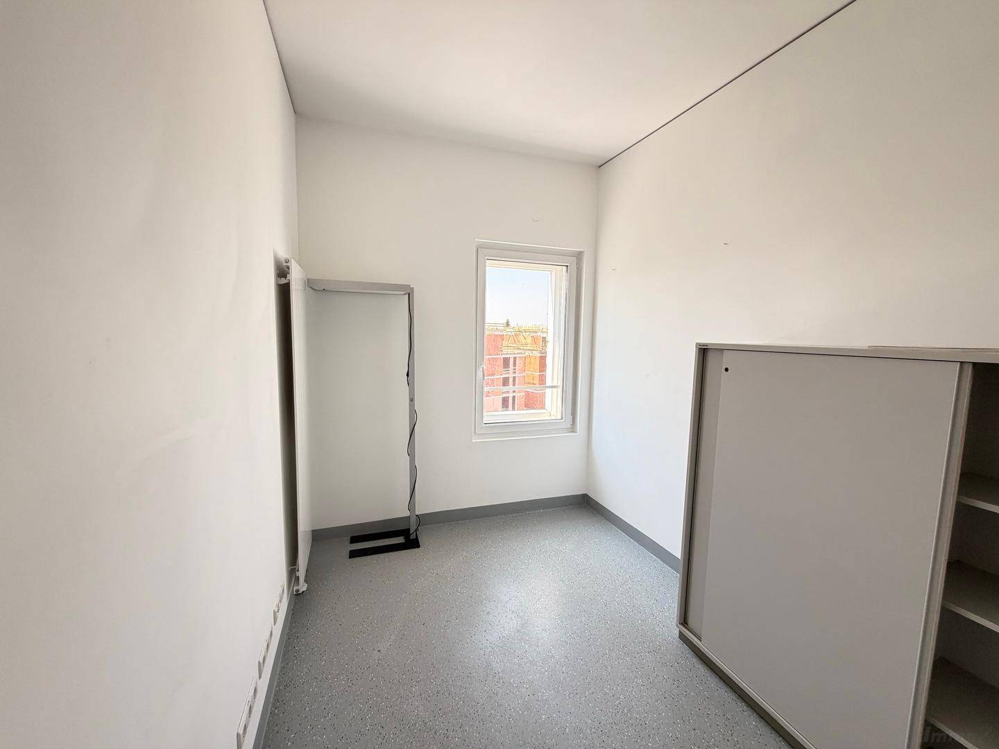 Schmaler Büroraum mit einem Fenster, weißen Wänden und einem praktischen Schrank an der Seite.