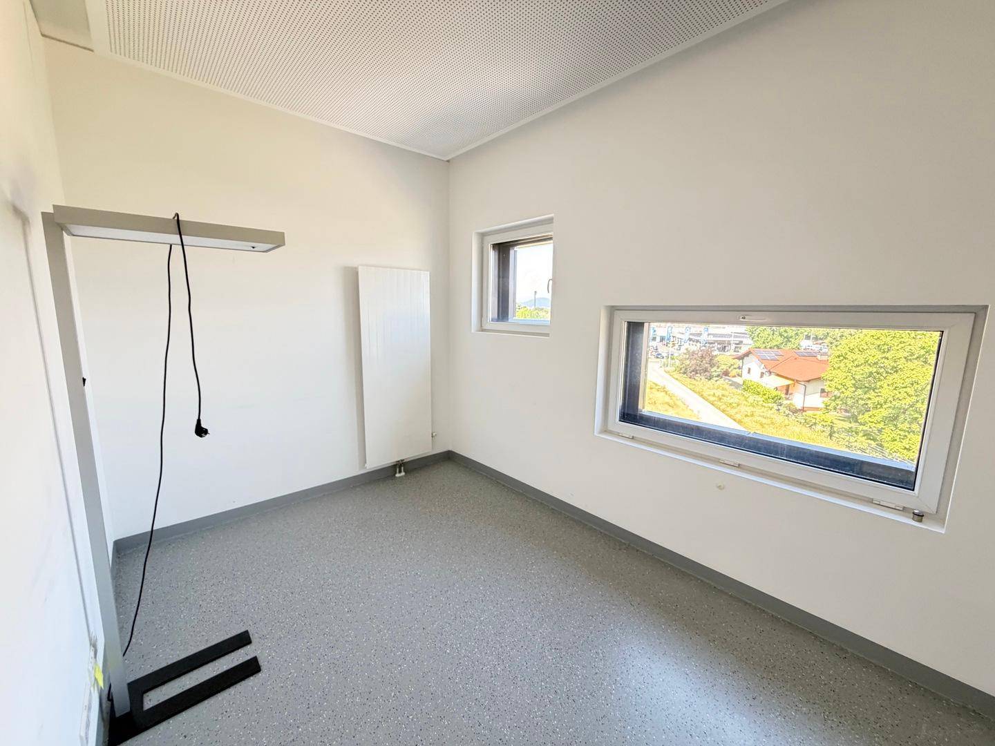 Heller Büroraum mit zwei Fenstern, grauen Böden und einem Heizkörper an der Wand, bereit zur Einrichtung.