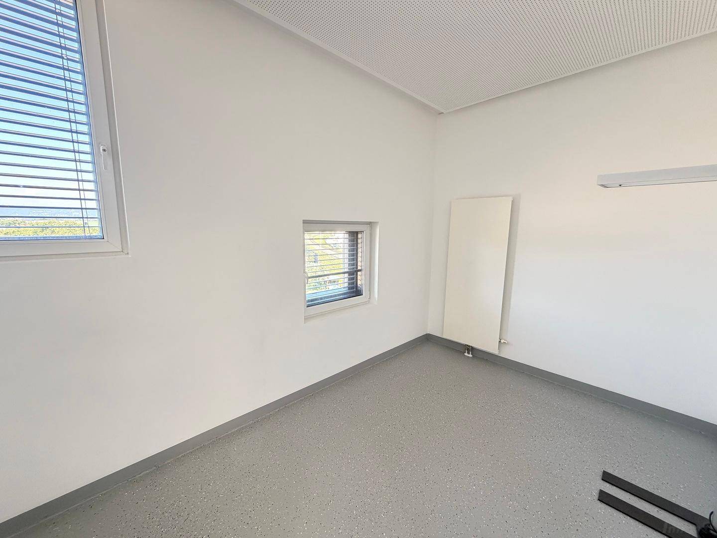 Leerer Büroraum mit grauen Vinylböden, weißen Wänden und Jalousien an den Fenstern für Privatsphäre.