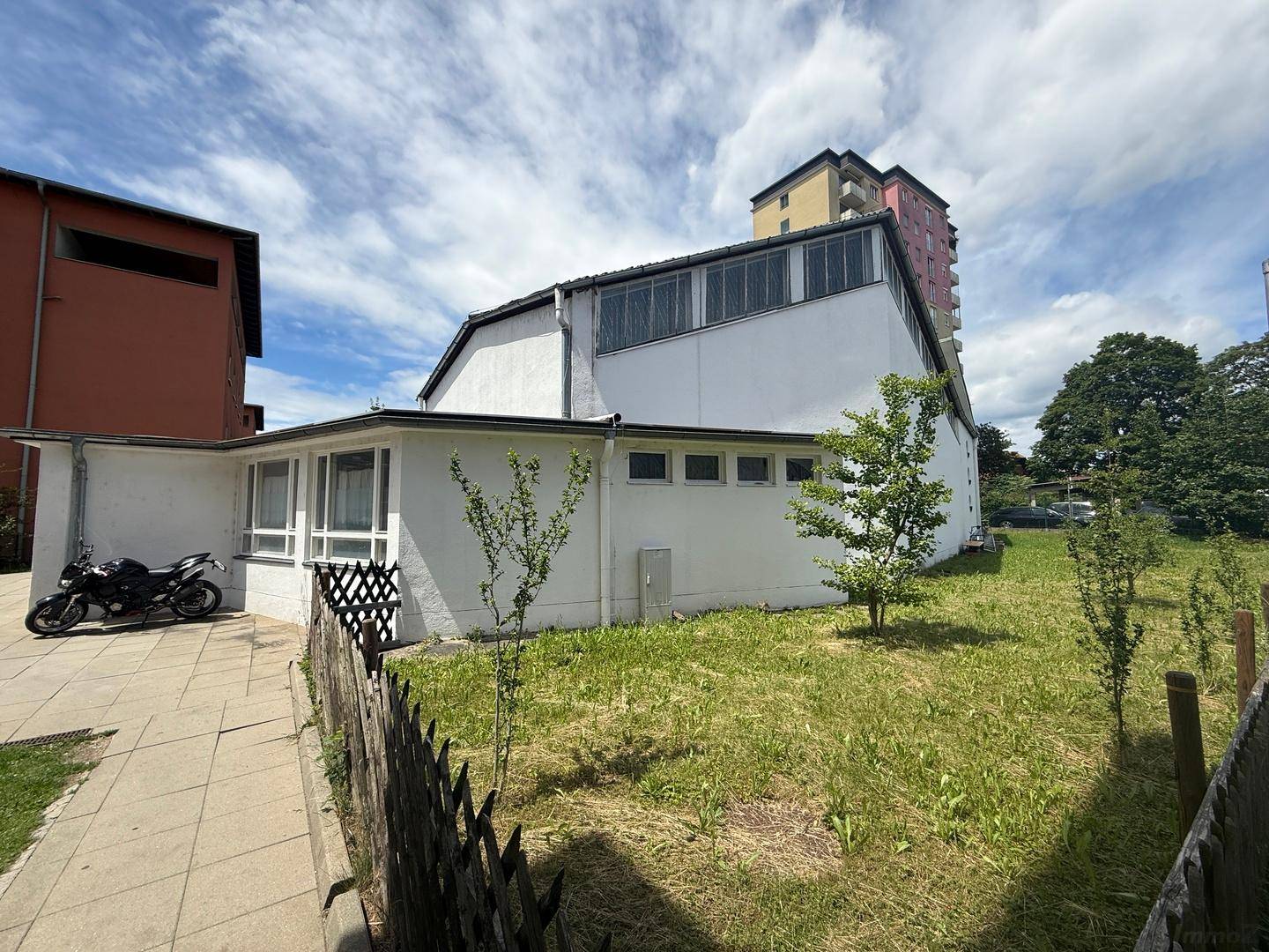 Seitenansicht des Gebäudes mit weißer Fassade, kleinen Bäumen und einem Motorrad auf dem Gehweg.