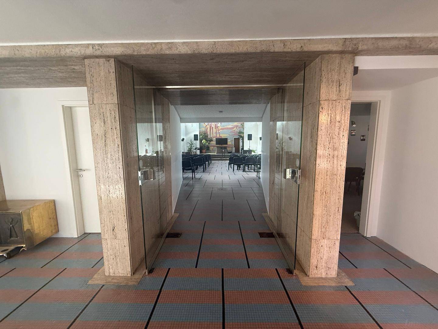 Blick durch Glastüren vom Flur in einen großen Saal mit Sitzreihen und Bühne.