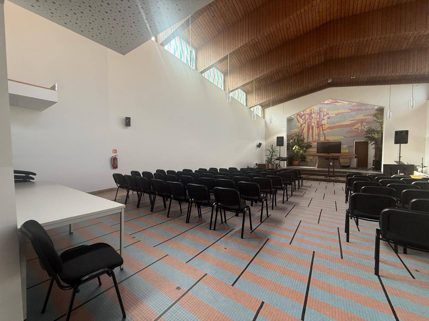Weitläufiger Saal mit Reihenbestuhlung, Holzdach und einer Bühne im Hintergrund, hell und geräumig.