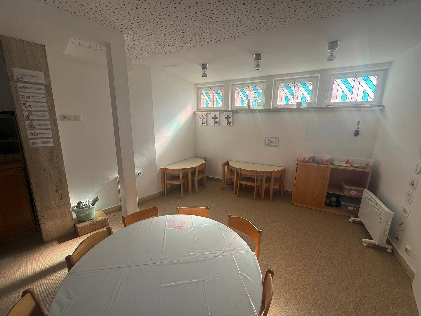 Freundlicher Raum mit Kindertischen, Teppichboden und farbigen Fenstern, ideal für Aktivitäten.