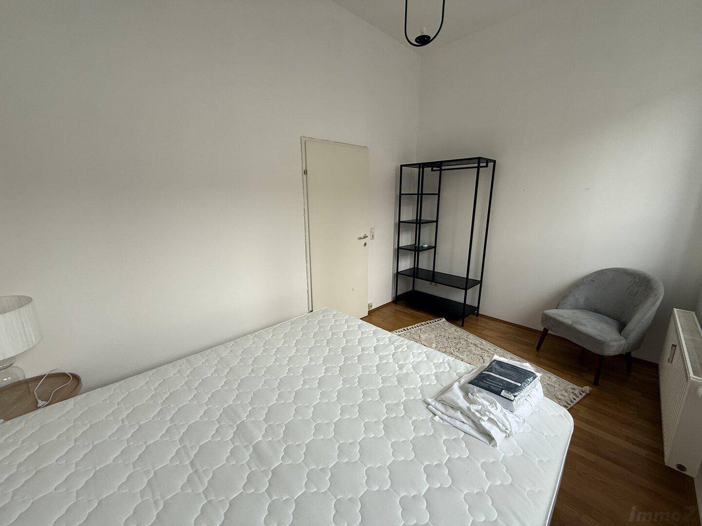 Schlafzimmer mit Doppelbett, offenem Kleiderschrank und minimalistischer Einrichtung.