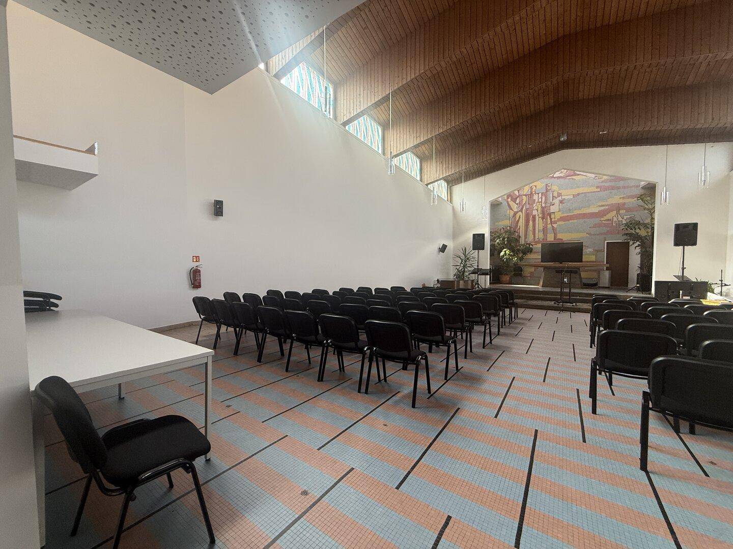 Geräumiger Saal mit vielen Stühlen, Holzdach und einem Wandgemälde im Hintergrund.