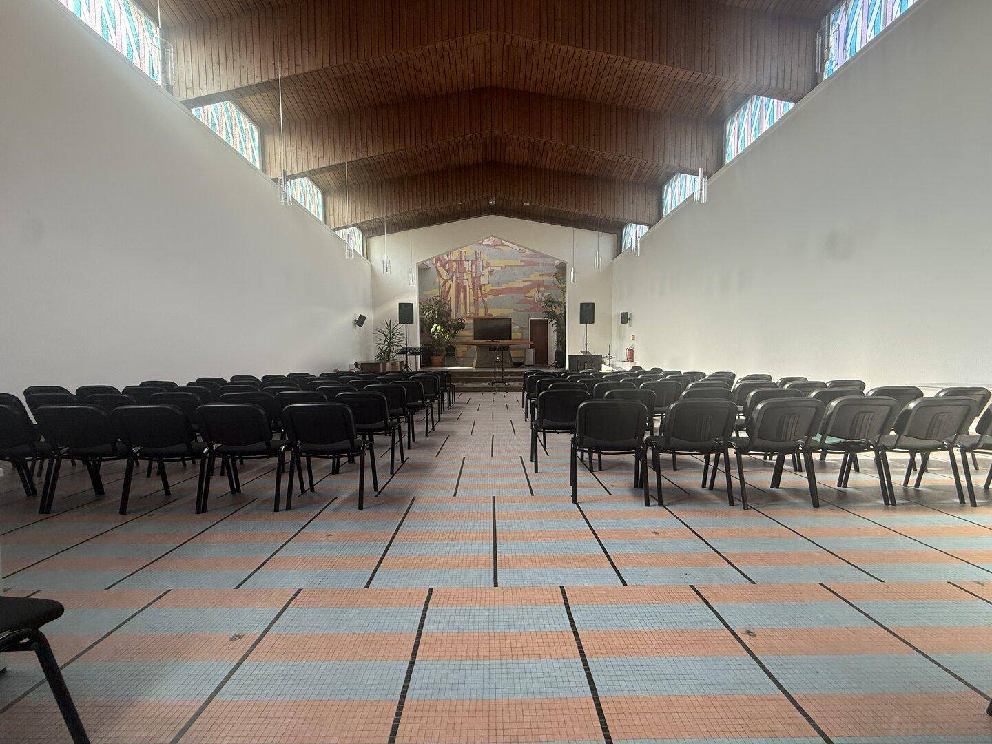 Großer Versammlungsraum mit Reihenbestuhlung, Holzdach und Wandgemälde.