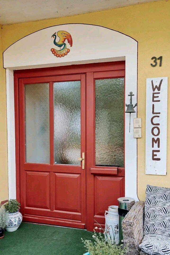 Rote Eingangstür mit Glaspaneelen und einer Willkommenstafel an der gelben Fassade.