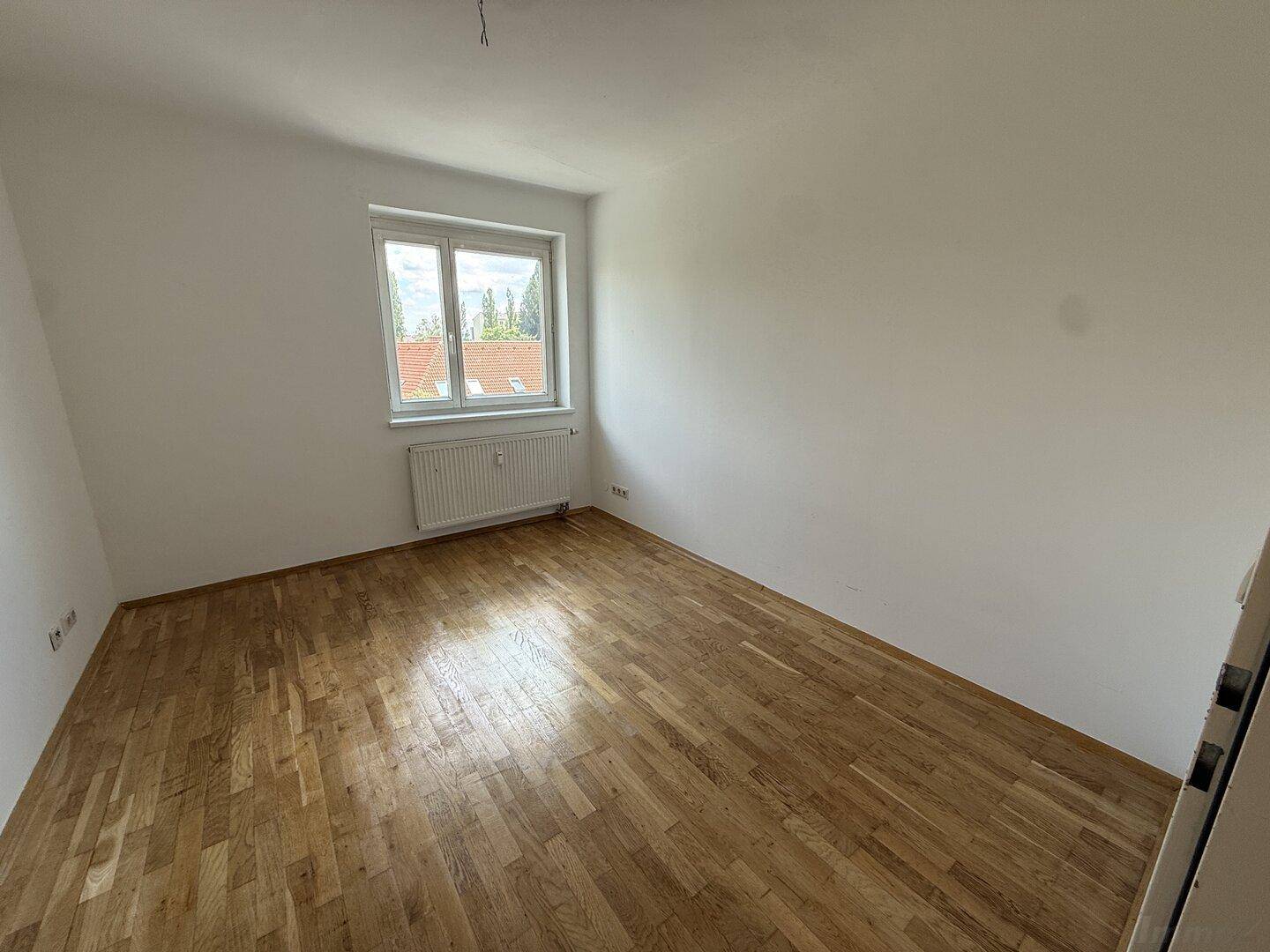 Leeres Zimmer mit Holzparkettboden und Fenster, das einen Blick ins Grüne bietet.