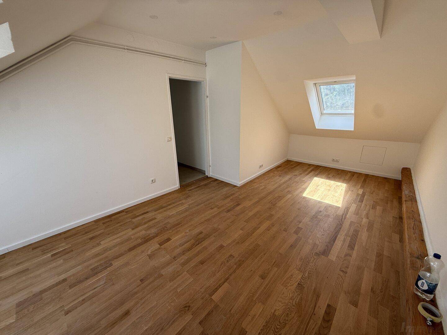 Dachgeschosszimmer mit Holzboden, Dachfenster und Zugang zu einem weiteren Bereich.