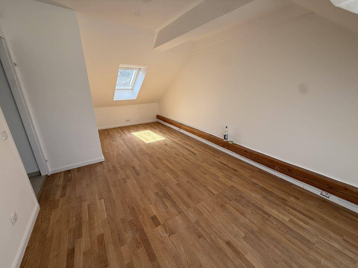 Helles Dachgeschosszimmer mit Holzboden und einem Dachfenster, das viel Sonnenlicht hereinlässt.