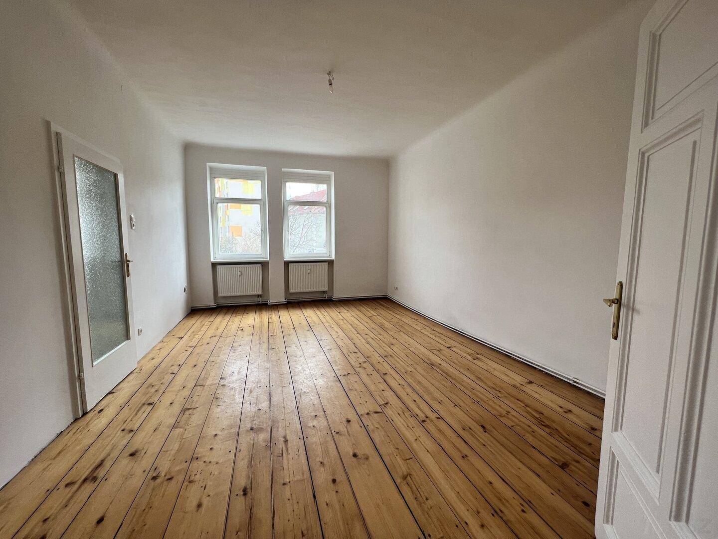 Helles Zimmer mit originalem Dielenboden, zwei Fenstern und weiß gestrichenen Wänden.