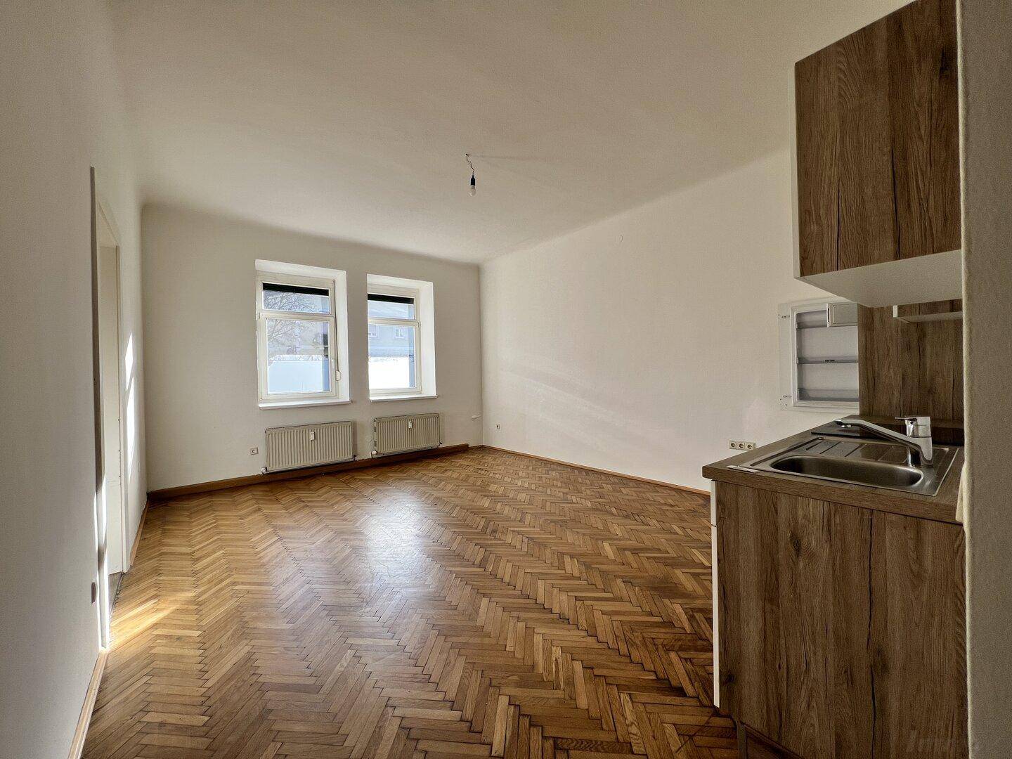 Leerer Raum mit Fischgrätparkett und einer kleinen Küchenzeile mit Spüle und Holzoptik-Schränken.