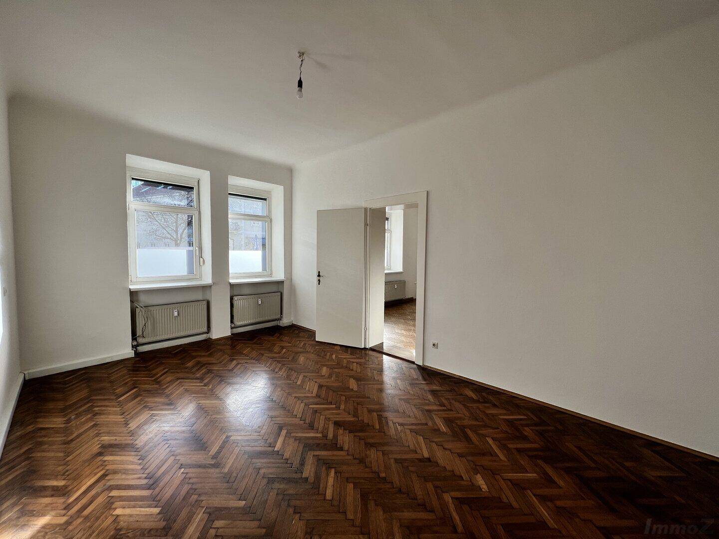 Geräumiges, helles Zimmer mit Fischgrätparkett und zwei Fenstern, das zu einem weiteren Raum führt.