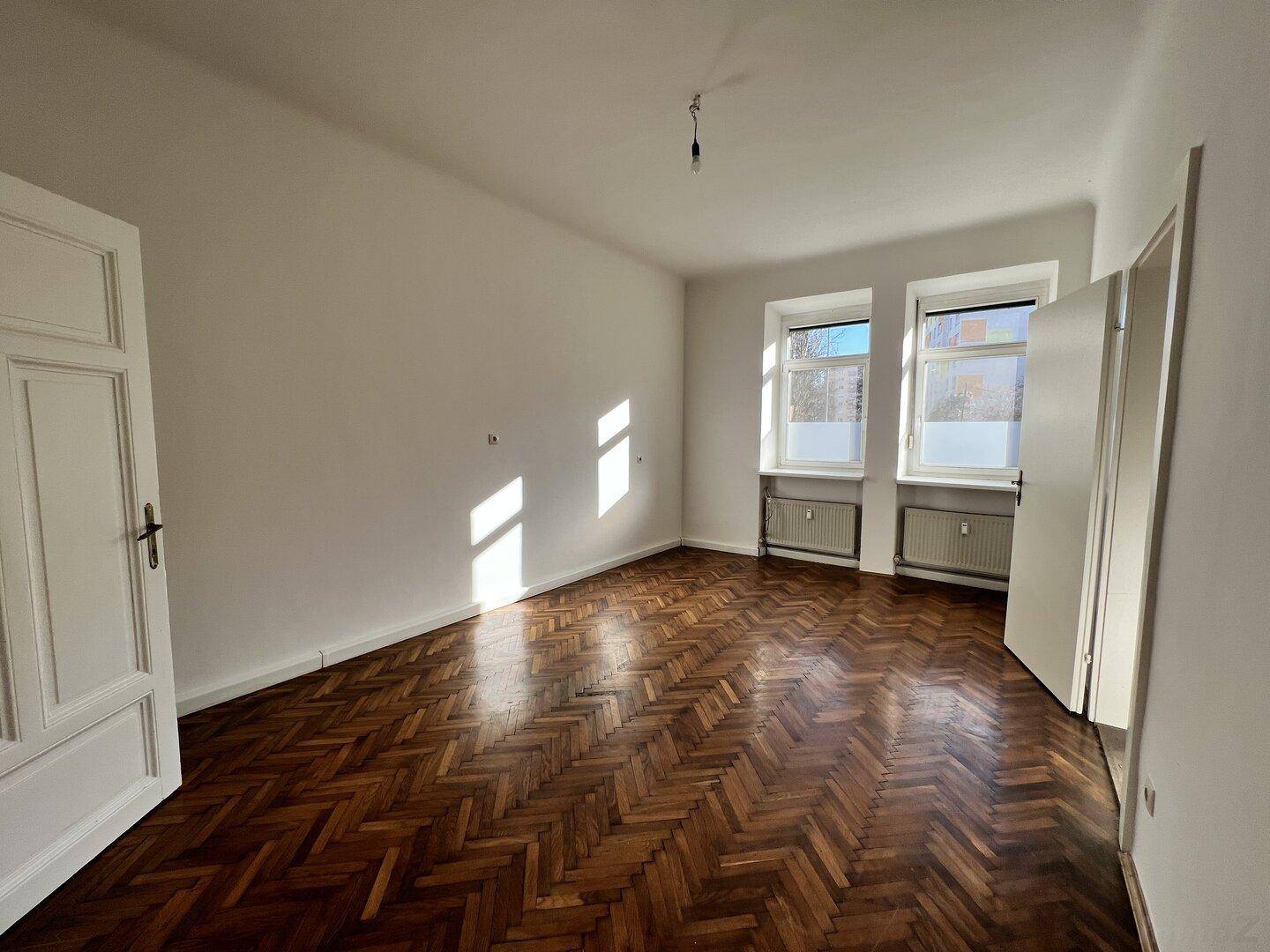 Lichtdurchflutetes Zimmer mit Fischgrätparkett und weißen Wänden, ideal für ein Wohn- oder Schlafzimmer.