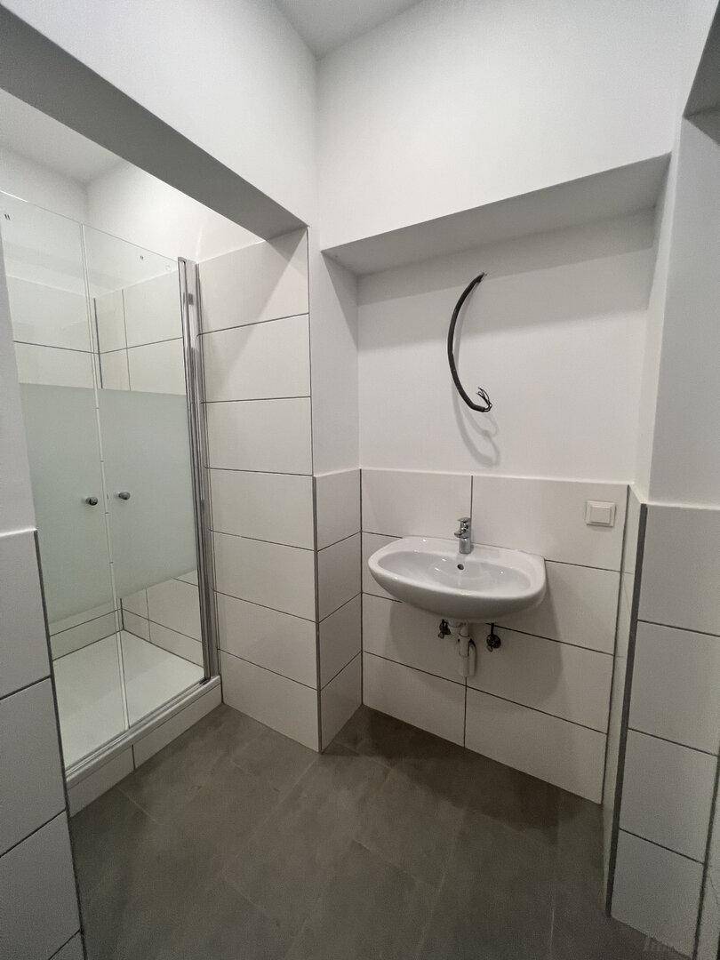 Helles Badezimmer mit grauen Bodenfliesen, weiß gefliesten Wänden und einer modernen Duschkabine.