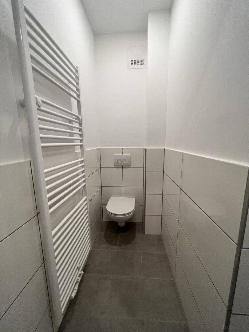 Separates WC mit wandmontierter Toilette, grauen Bodenfliesen und einem beheizten Handtuchhalter.