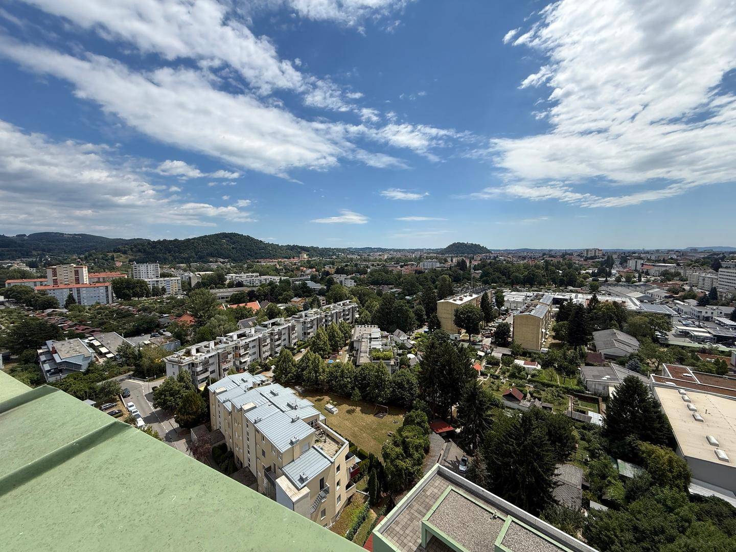Weitläufiger Ausblick über die Stadt mit Wohngebäuden, viel Grün und hügeliger Landschaft im Hintergrund.