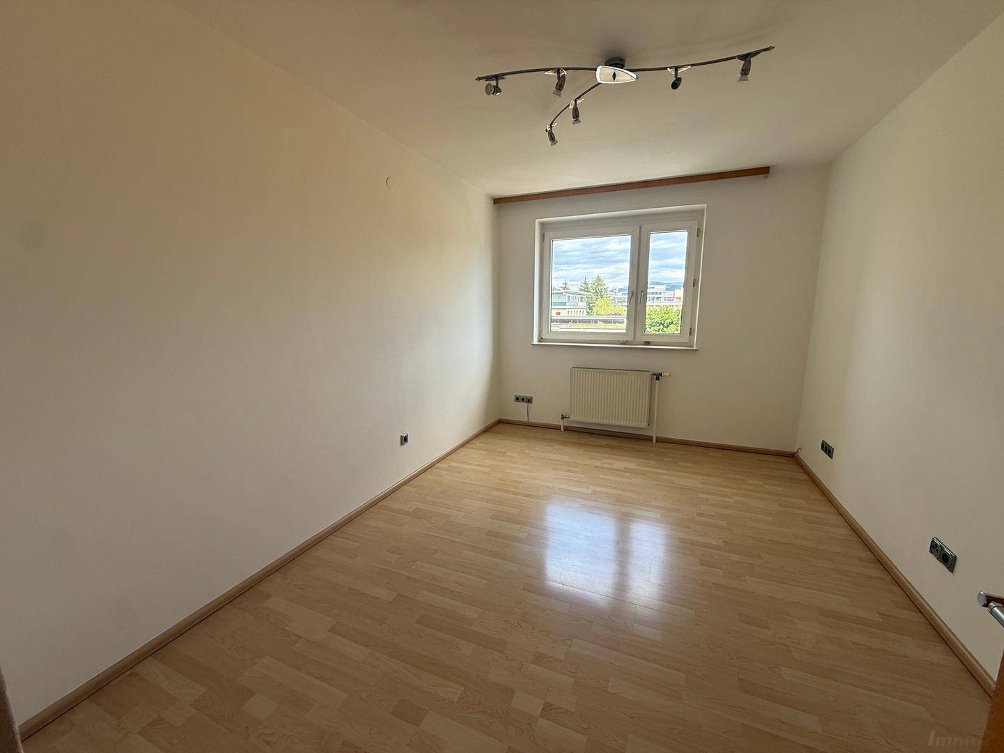 Leeres Zimmer mit hellem Holzfußboden, weißen Wänden und einem Fenster mit Blick ins Grüne.