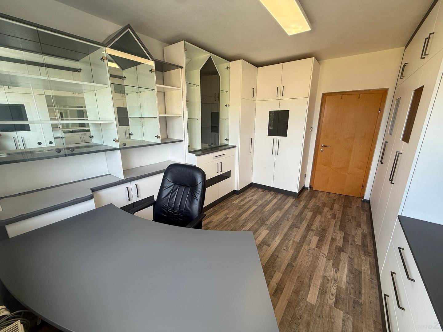 Funktionales Büro mit großem Schreibtisch, Bürostuhl und vielen weißen Einbauschränken mit Glasfronten.