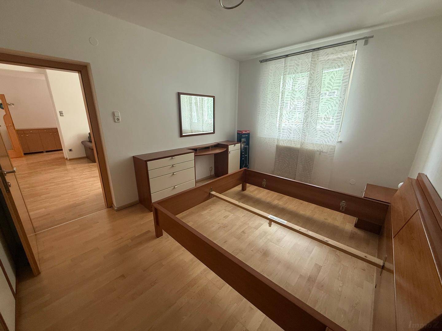 Helles Schlafzimmer mit Holzbett, Kommode und Fenster mit Vorhängen für Privatsphäre.