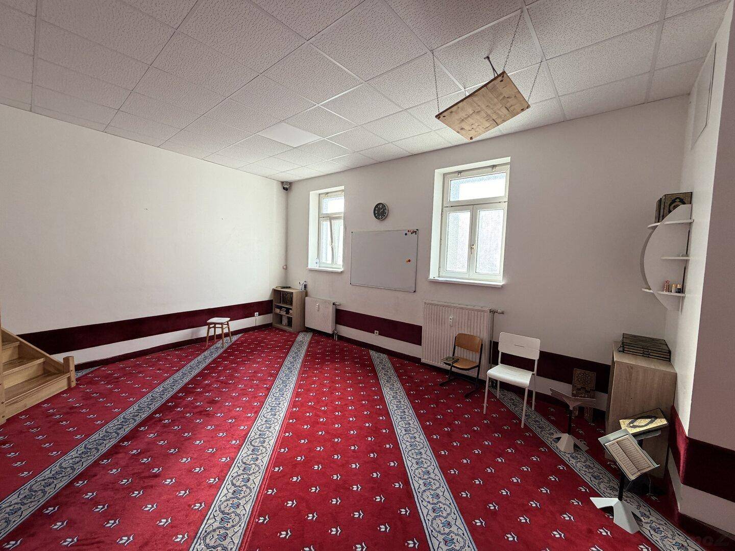 Großer Raum mit rotem Teppichboden, weißen Wänden und zwei Fenstern, ausgestattet mit einer Holztreppe.