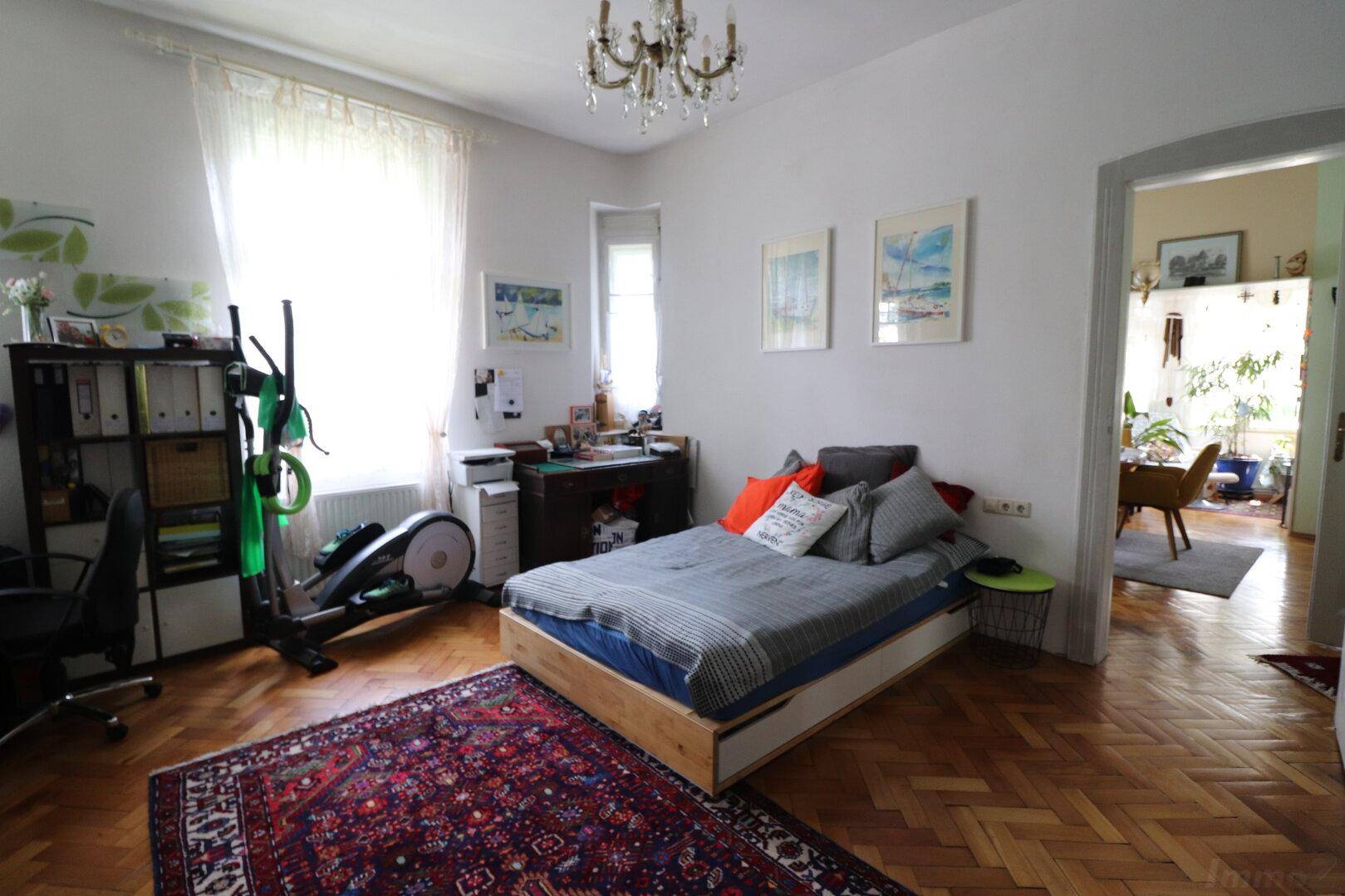 Helles Schlafzimmer mit Parkettboden, Bett und Fitnessgerät, verbunden mit einem weiteren Raum.