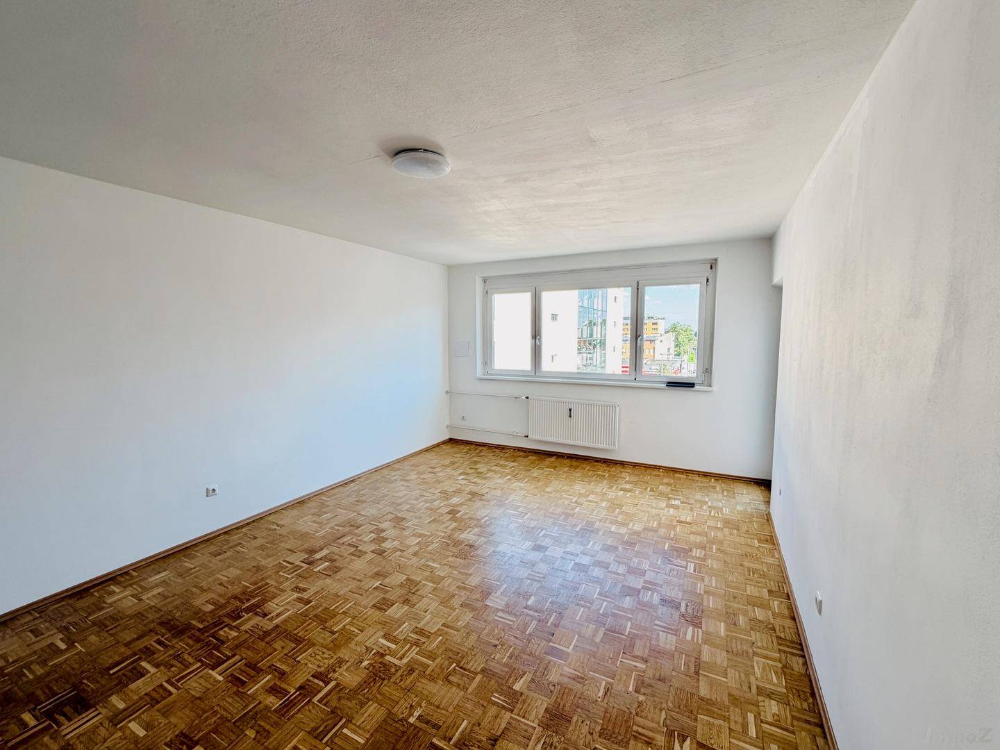 Geräumiges Zimmer mit hellem Fischgrätparkett und großen Fenstern, ideal als Wohn- oder Schlafzimmer.