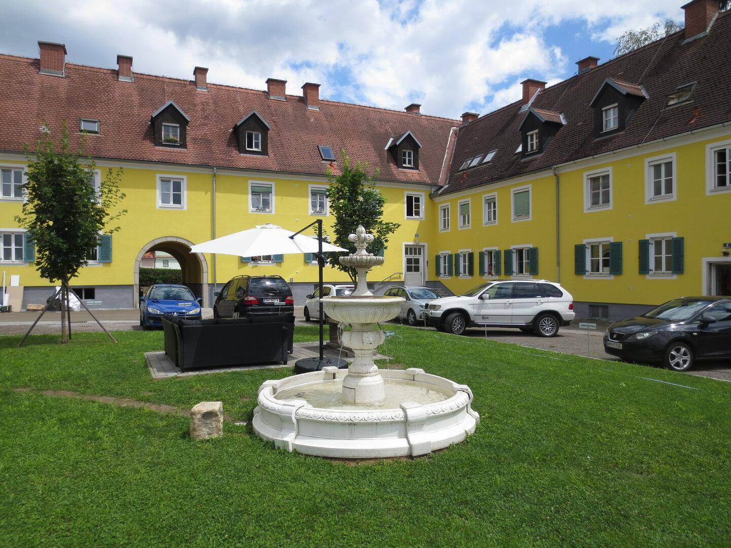 Innenhof mit einem dekorativen Brunnen, Rasen und geparkten Autos, umgeben von gelben Gebäuden.