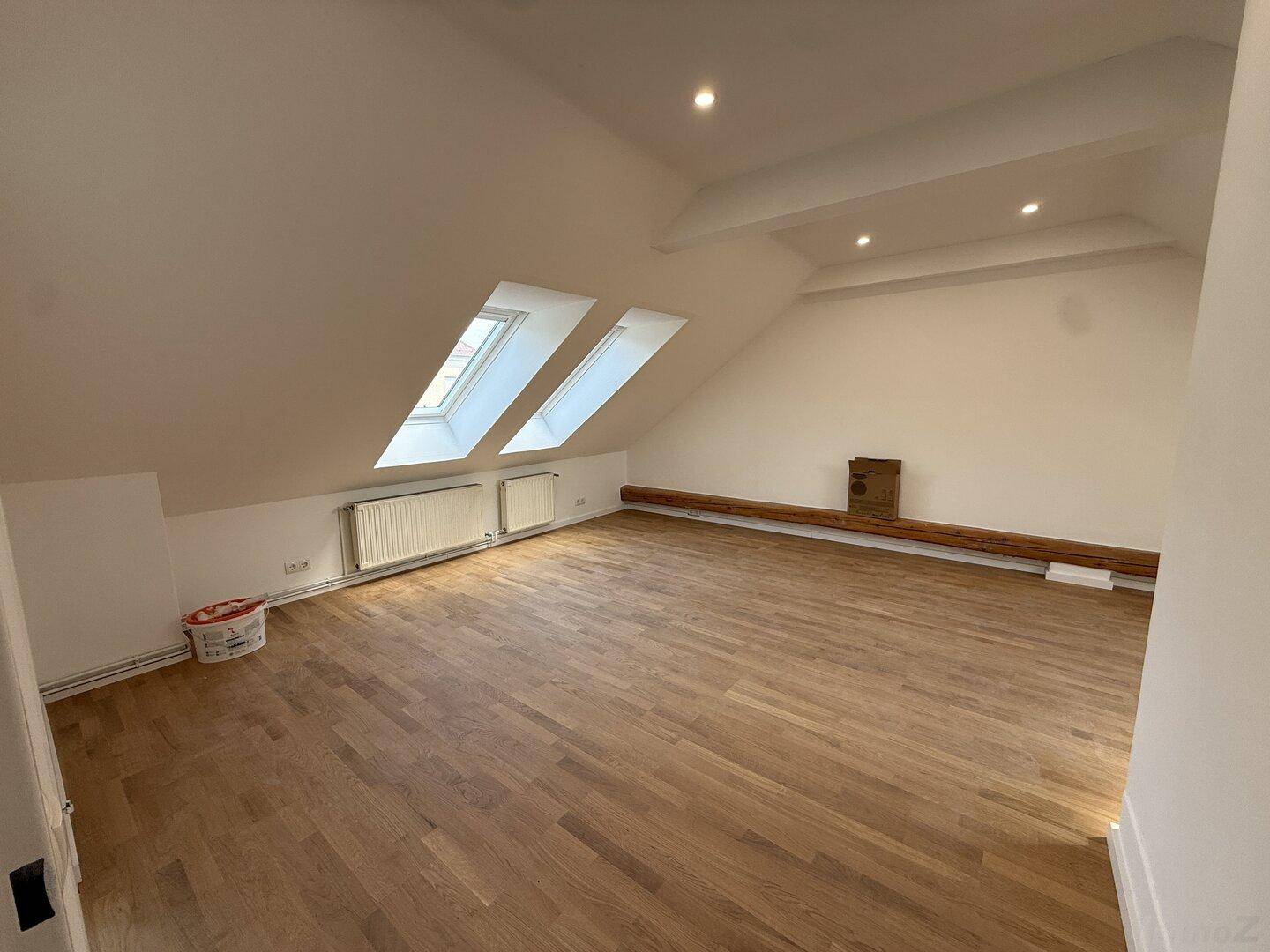 Großer Dachgeschossraum mit Holzboden, zwei Dachfenstern und Heizkörpern.