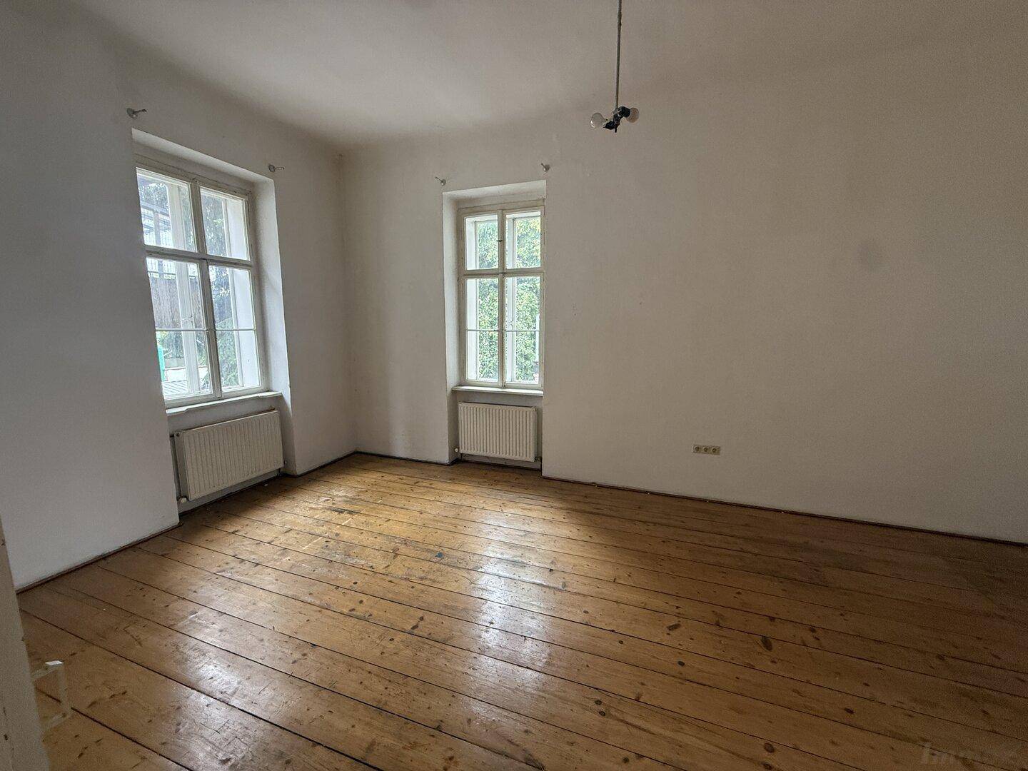 Geräumiges Zimmer mit Holzdielenboden und zwei Fenstern, ideal für Wohn- oder Schlafbereich.