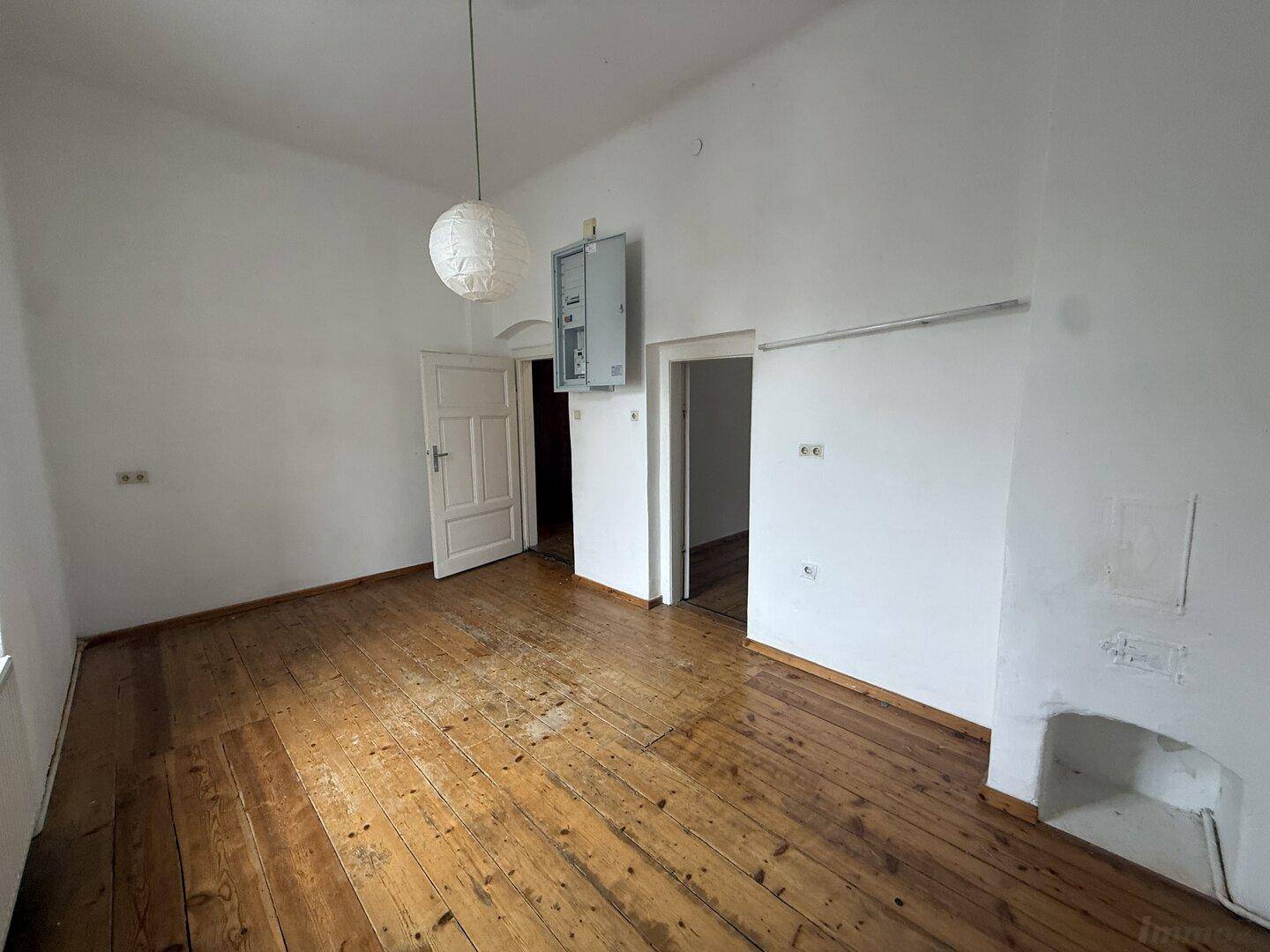 Zimmer mit Holzdielenboden, Sicherungskasten und Zugang zu weiteren Räumen, bereit zur Renovierung.