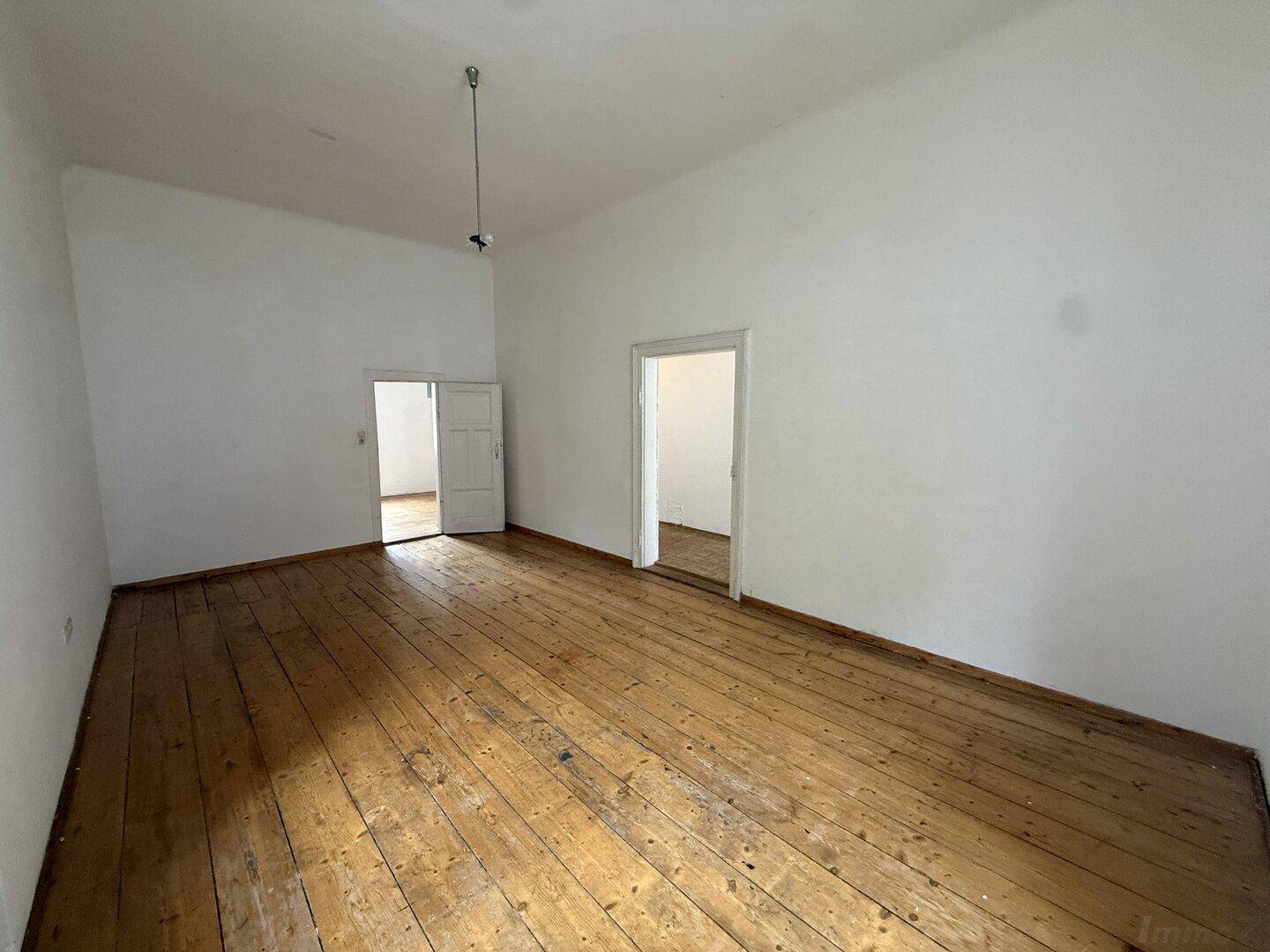 Großes, leeres Zimmer mit Holzdielenboden und zwei offenen Türen zu angrenzenden Räumen.
