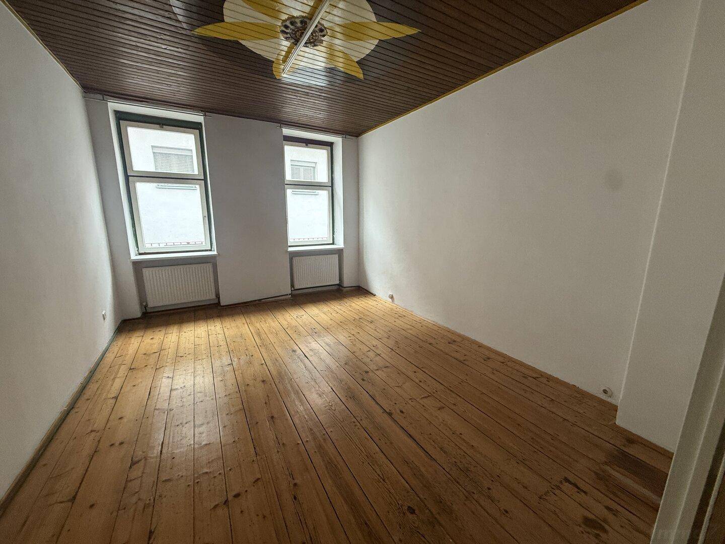 Zimmer mit Holzdielenboden und Holzdecke, zwei Fenstern und dekorativer Deckenleuchte.