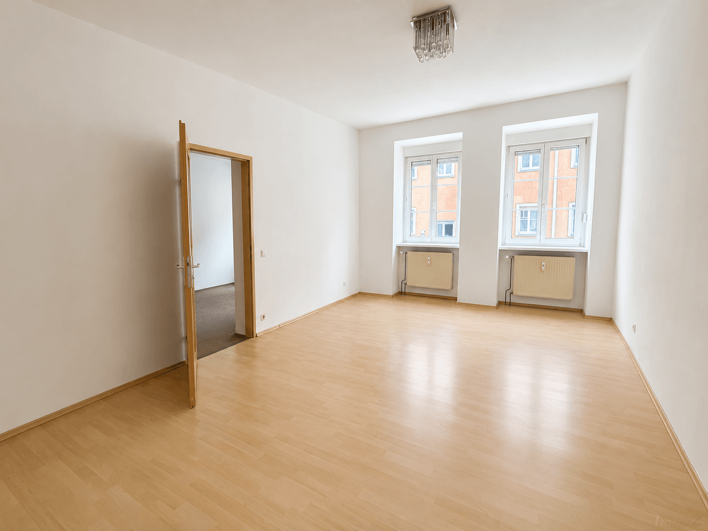 Helles, leeres Wohnzimmer mit Laminatboden und zwei Fenstern, die viel Tageslicht hereinlassen.