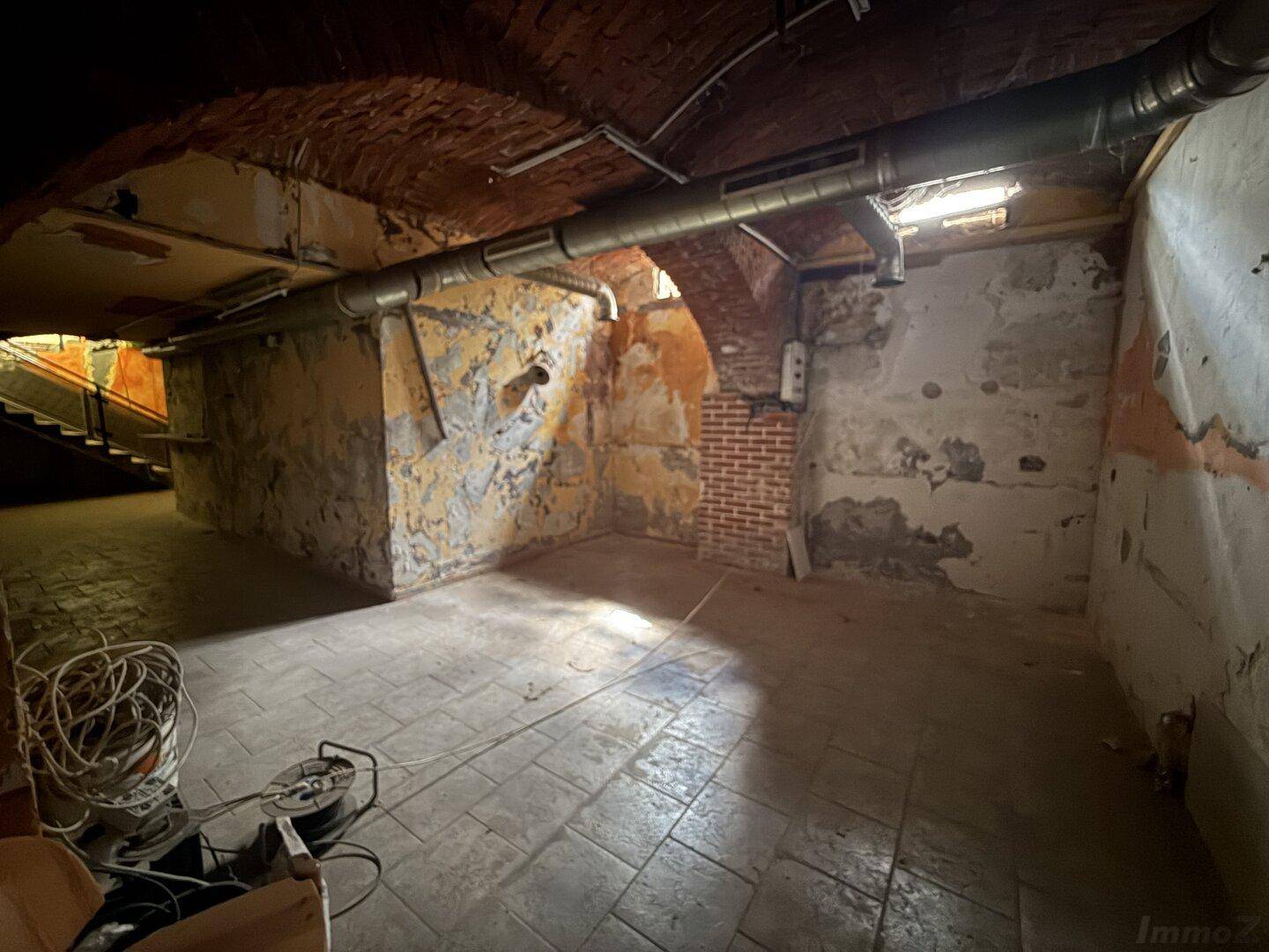 Weiterer Blick in den rohen Kellerraum mit Ziegelgewölbe, freiliegenden Rohren und stark beschädigten Wänden.