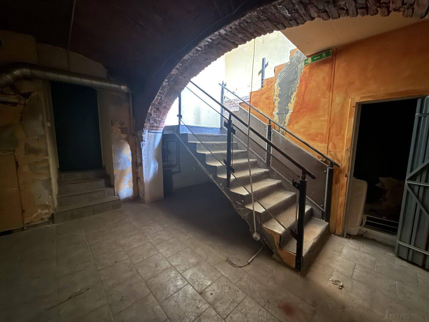 Treppenhaus im Keller mit Metalltreppe und Geländer, das unter einem Ziegelbogen nach oben führt.
