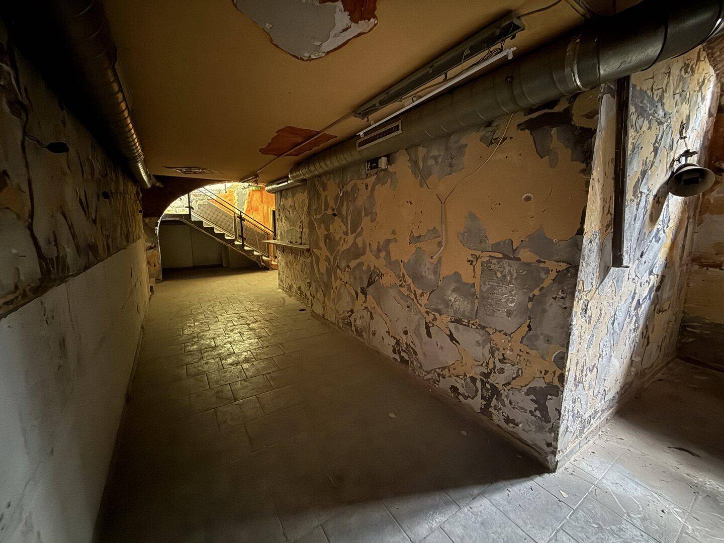 Langer Flur im Keller mit Ziegelgewölbedecke, sichtbaren Rohren und abblätternder Wandfarbe, Treppe im Hintergrund.