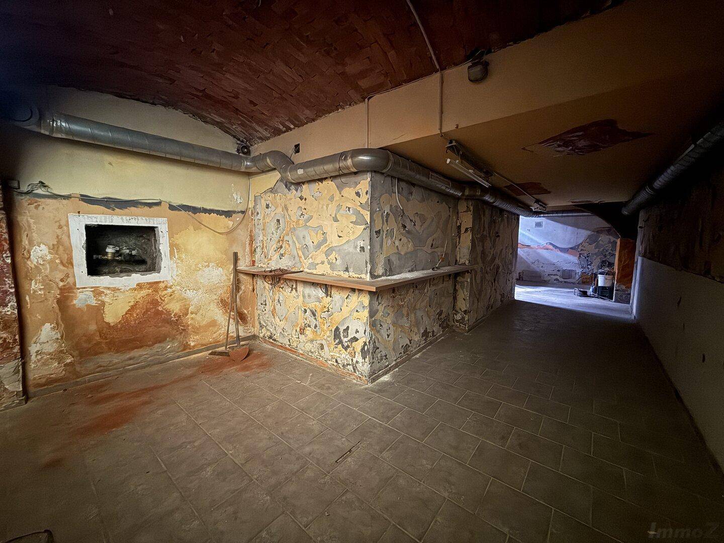 Ecke eines rohen Kellerraums mit beschädigten Wänden, Ziegelgewölbedecke und freiliegenden Lüftungsrohren.