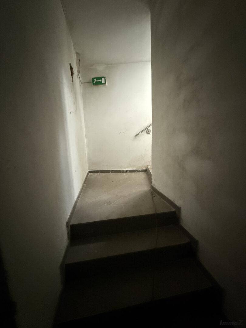 Dunkles Treppenhaus mit weißen Wänden, Betonstufen und einem Handlauf, ein Notausgangsschild ist sichtbar.