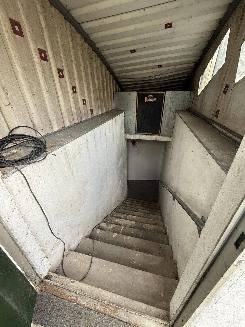 Treppenhaus führt in einen Keller hinunter, mit Betonwänden und Stufen, ein Safe ist unten sichtbar.