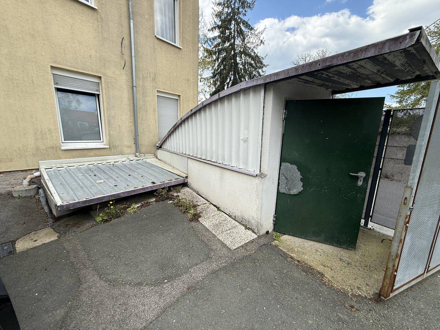 Außenansicht eines Gebäudes mit überdachtem Eingang, grüner Tür und einer Rampe oder Plattform.