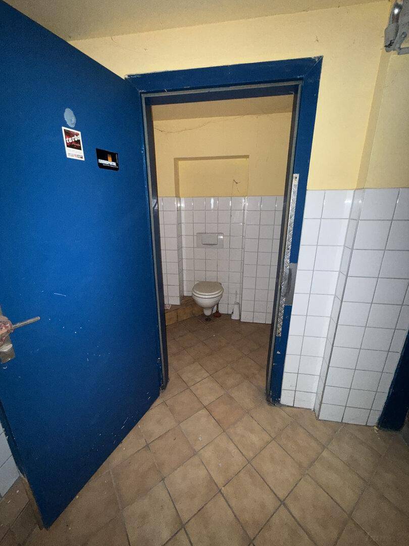 Blick durch eine blaue Tür in eine Toilettenkabine mit WC, gefliesten Wänden und Fliesenboden.