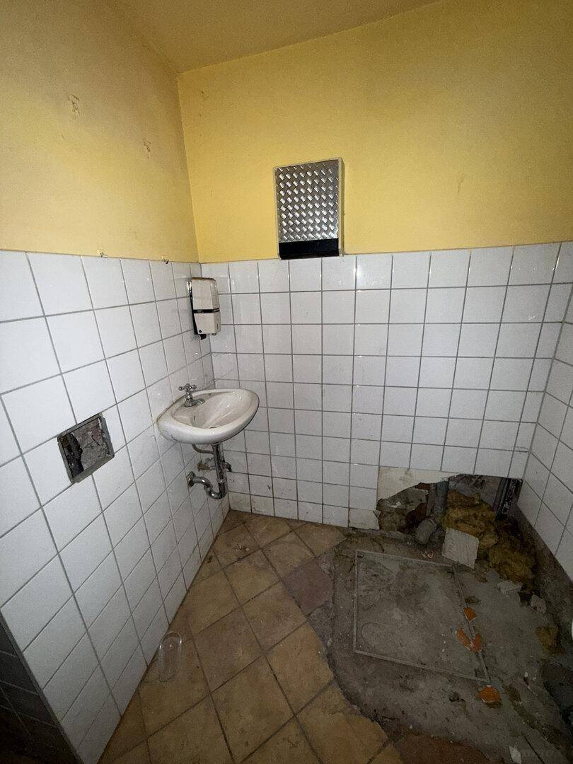 Weiterer Blick in die alte öffentliche Toilette mit Waschbecken, gefliesten Wänden und beschädigtem Boden.