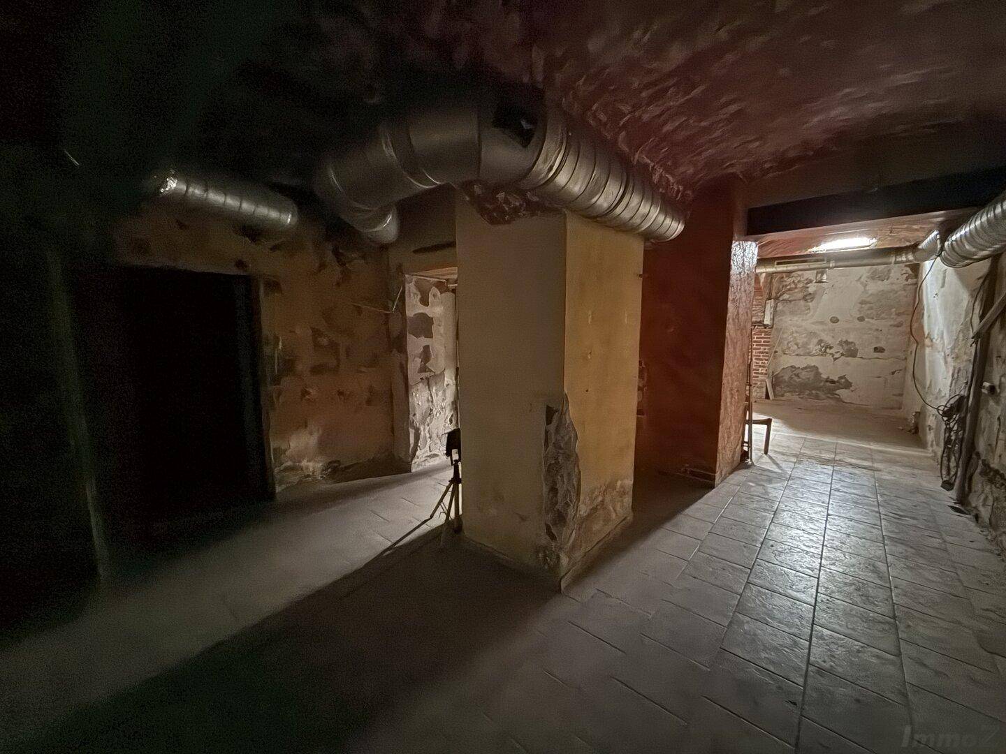 Weiterer dunkler Kellerraum mit freiliegenden Rohren, beschädigten Wänden und einem tragenden Pfeiler.
