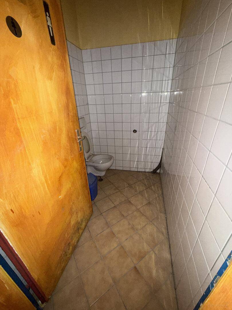 Toilettenkabine mit WC, weißen Fliesen an den Wänden und braunen Fliesen auf dem Boden, Holztür.