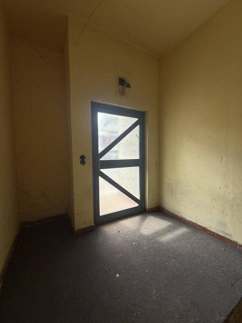 Kleiner, alter Raum mit gelben Wänden, dunklem Boden und einer blaugrauen Tür, durch die Licht fällt.