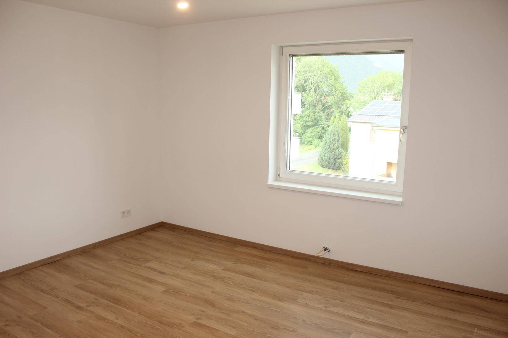 Helles, leeres Zimmer mit Laminatboden und einem Fenster, das einen Blick ins Grüne bietet.