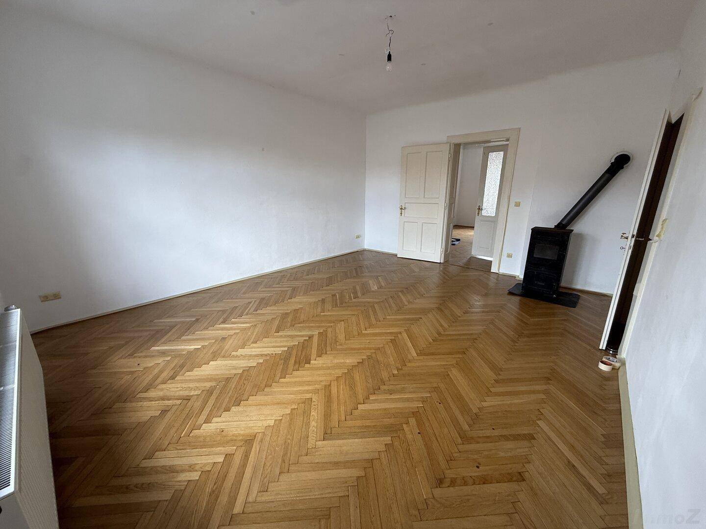 Geräumiges Zimmer mit Fischgrätparkett, weiß gestrichenen Wänden und einem gemütlichen Holzofen.