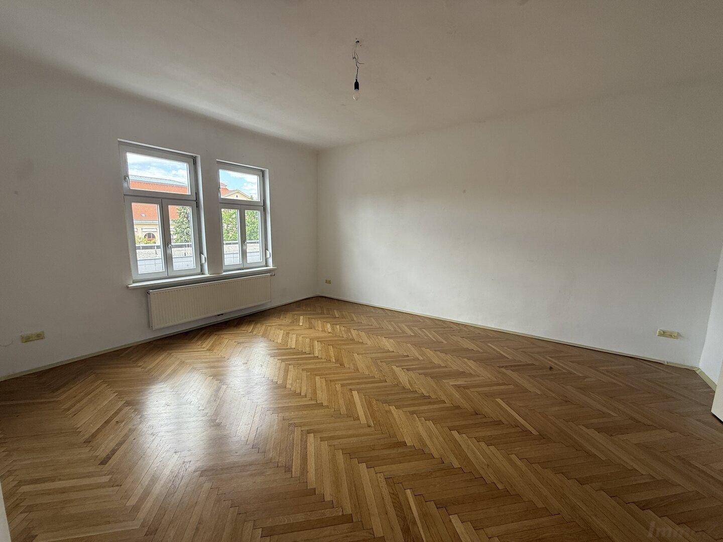 Großes, helles Zimmer mit Fischgrätparkett und zwei Fenstern, ideal für Wohn- oder Schlafbereich.