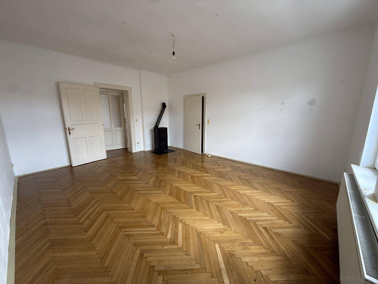 Helles Zimmer mit Fischgrätparkett, zwei Türen und einem freistehenden Holzofen als Wärmequelle.